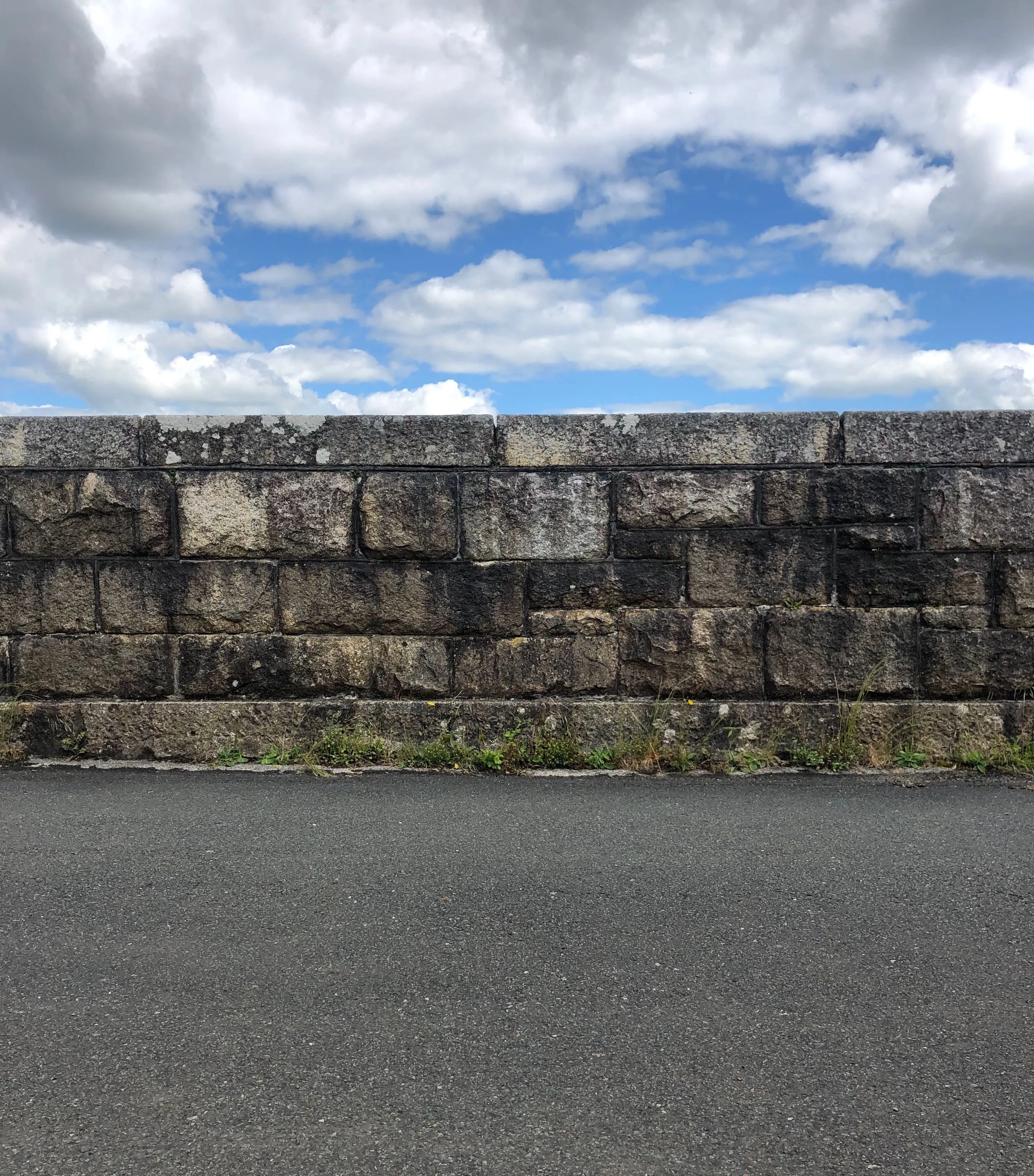 Photography of a stone wall running alongside a path. A bright blue sky and bright clouds are visible in the background.